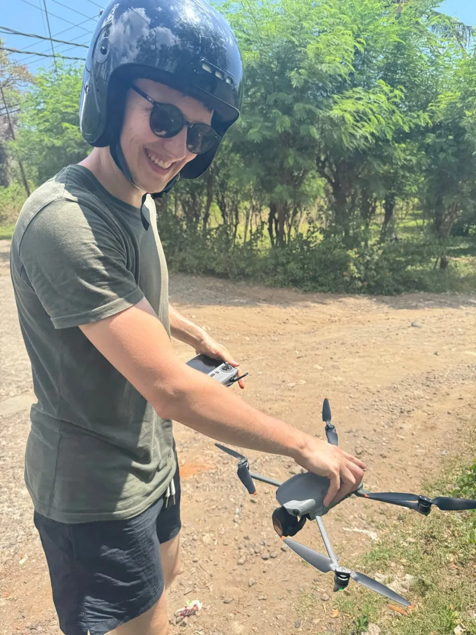 Ein lächelnder Mann mit Helm und Sonnenbrille hält auf einem Feldweg eine Drohne und die Fernsteuerung in den Händen