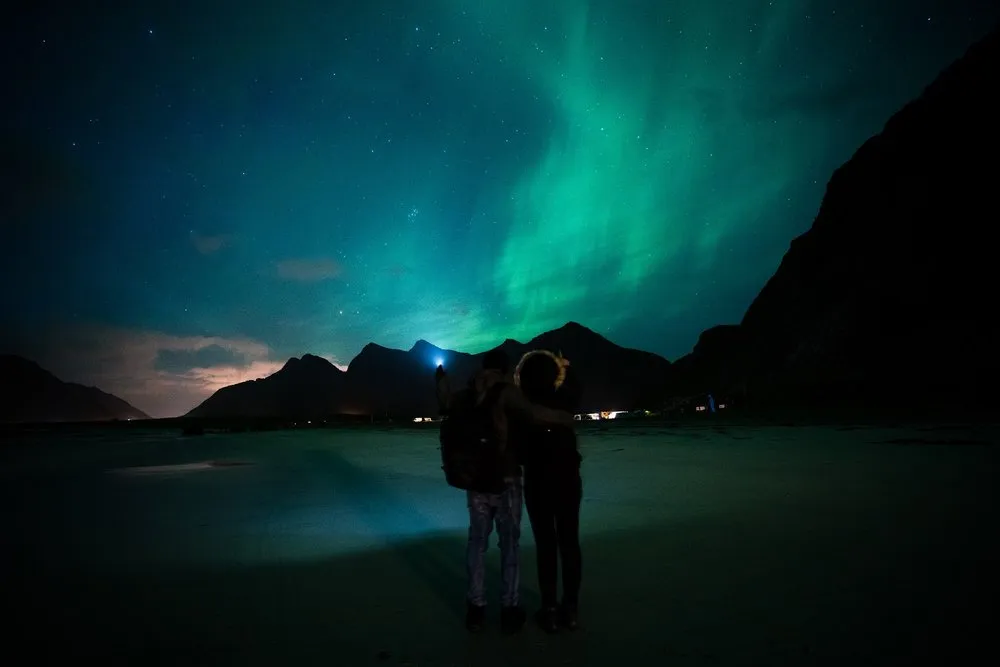 Zwei Personen stehen nachts eng beieinander an einem Sandstrand, über den Bergen leuchten grüne Nordlichter