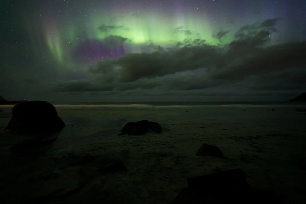 Green and violet northern lights above dark clouds, a beach with boulders below