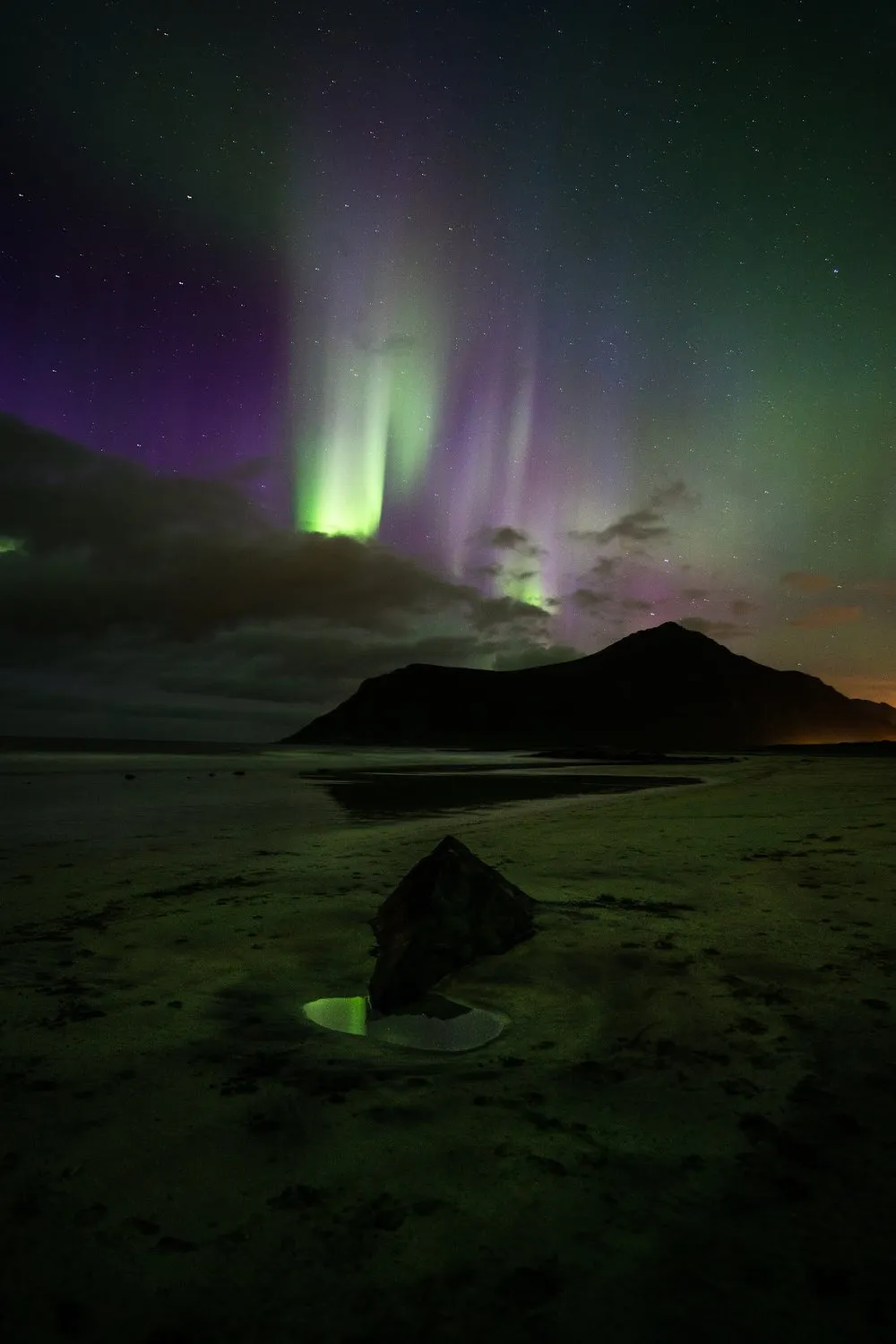 Violet and green streaks of light above a dark mountain, in the foreground a rock and a puddle in the sand reflecting green