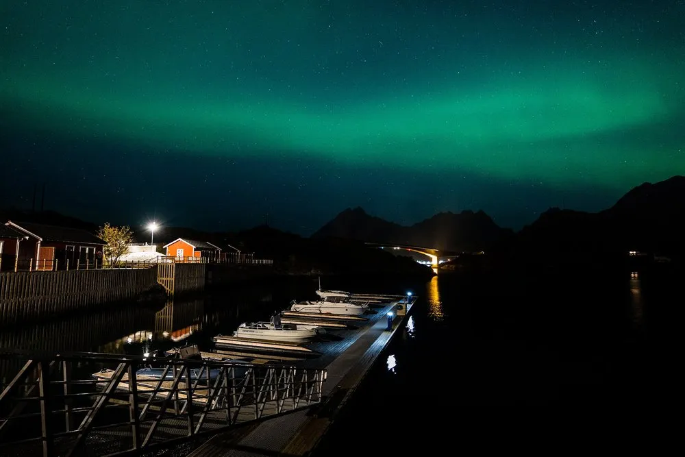 Green northern lights above a small harbour with boats at the jetty, red houses and a bridge in the background