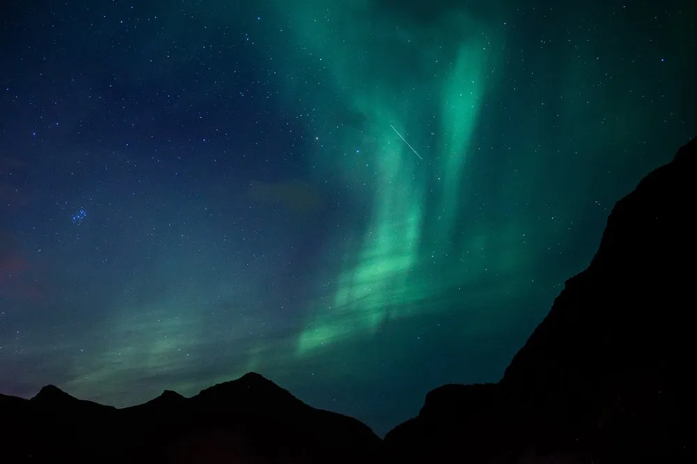 Green northern lights rise behind black mountain silhouettes into the starry sky