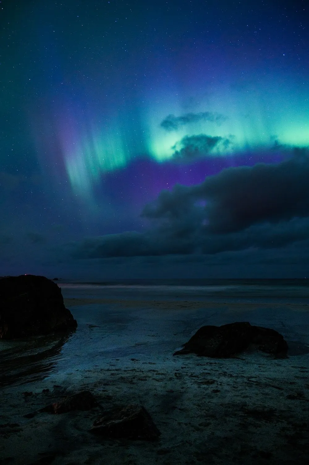 Green and blue glowing streaks above dark clouds, rocks and wet sand on the beach below