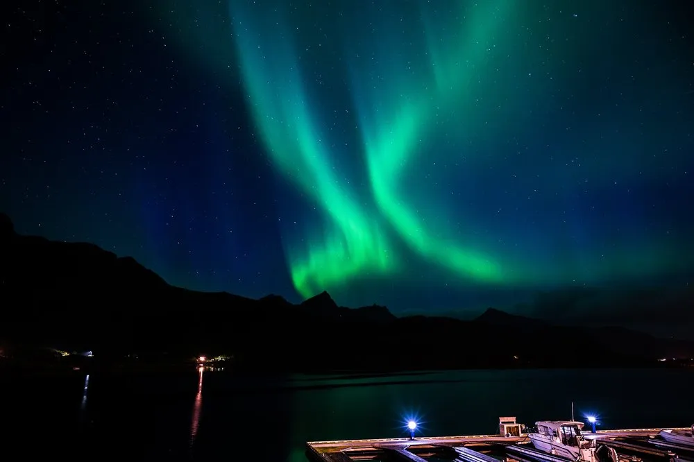 Green ribbons of light above the water, at the bottom right a jetty with boats and blue lamps