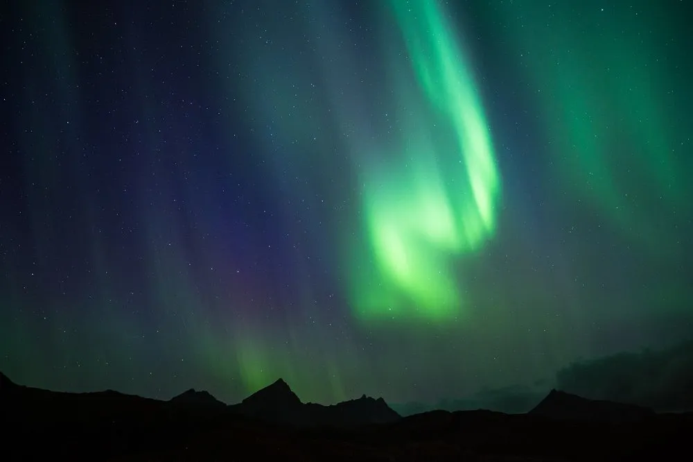 A northern light with green and violet rays above the peaks of dark mountains