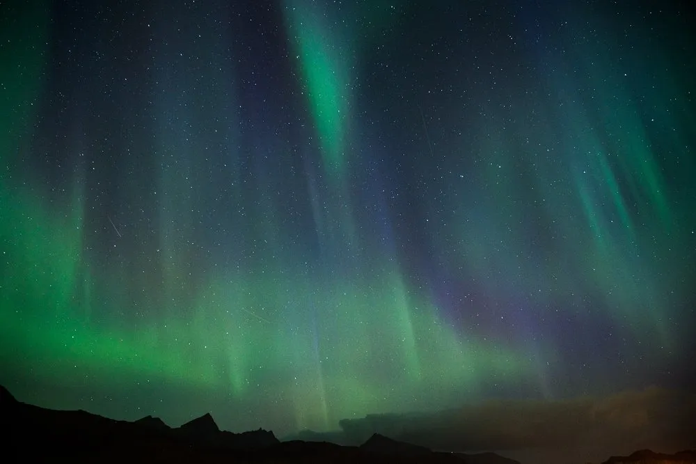 Vertical streaks of light in green and violet against many stars above a mountain range