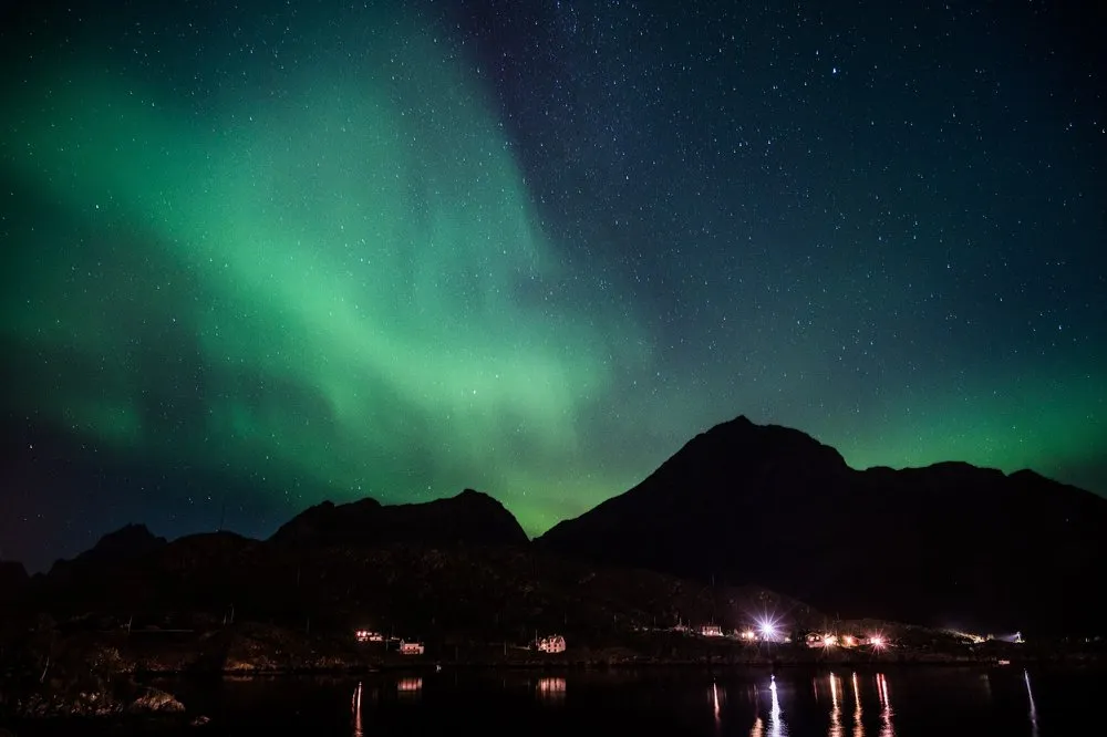 Broad green veils of light drift diagonally across a mountain, lights burn on the shore