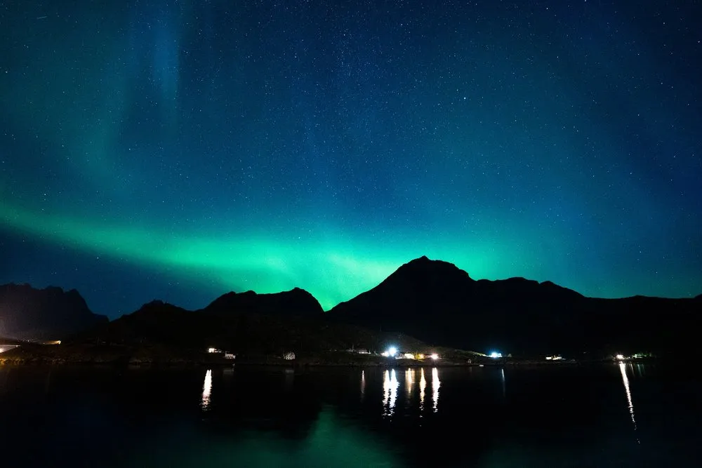 A green ribbon of light behind a mountain, the lights on the shore reflected in the calm water