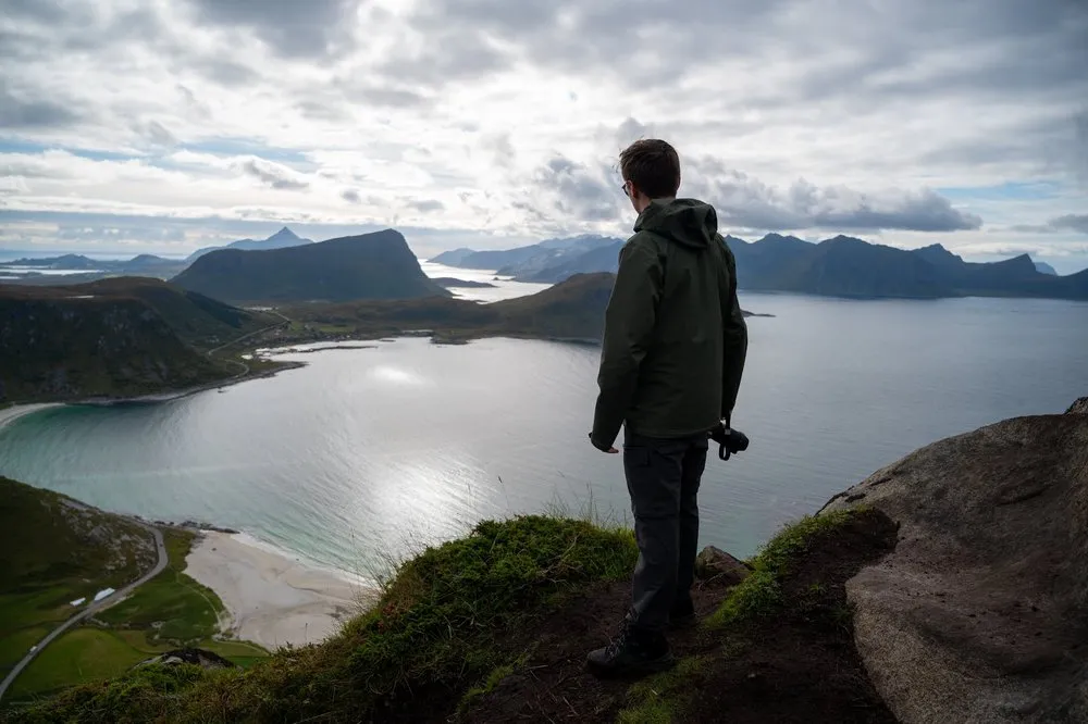 A man in a green jacket stands at the edge with a camera in his hand, looking at the bay and mountains