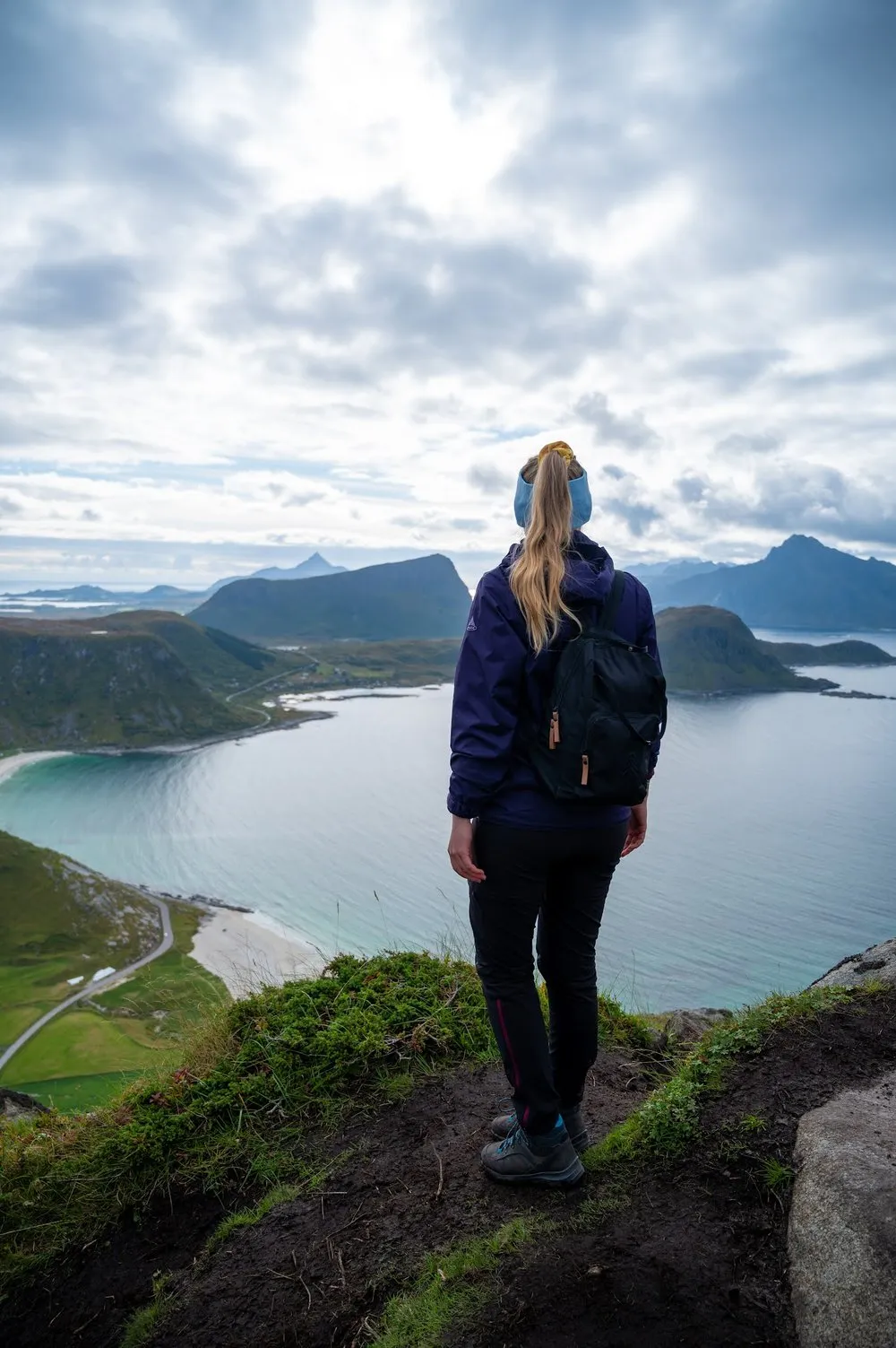 A woman with a backpack and a blue headband stands at the edge looking at the bay and mountains