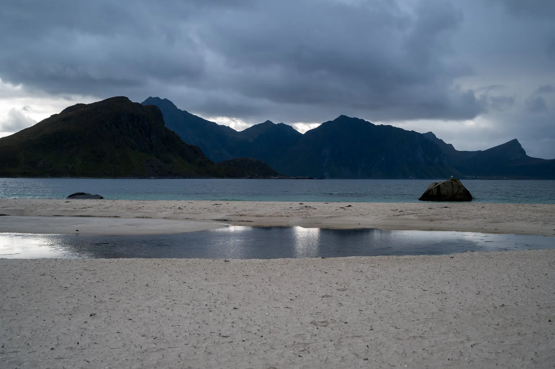 Am Hauklandstranda spiegelt eine Pfütze im hellen Sand den Himmel, dahinter ein Felsblock und dunkle Berge