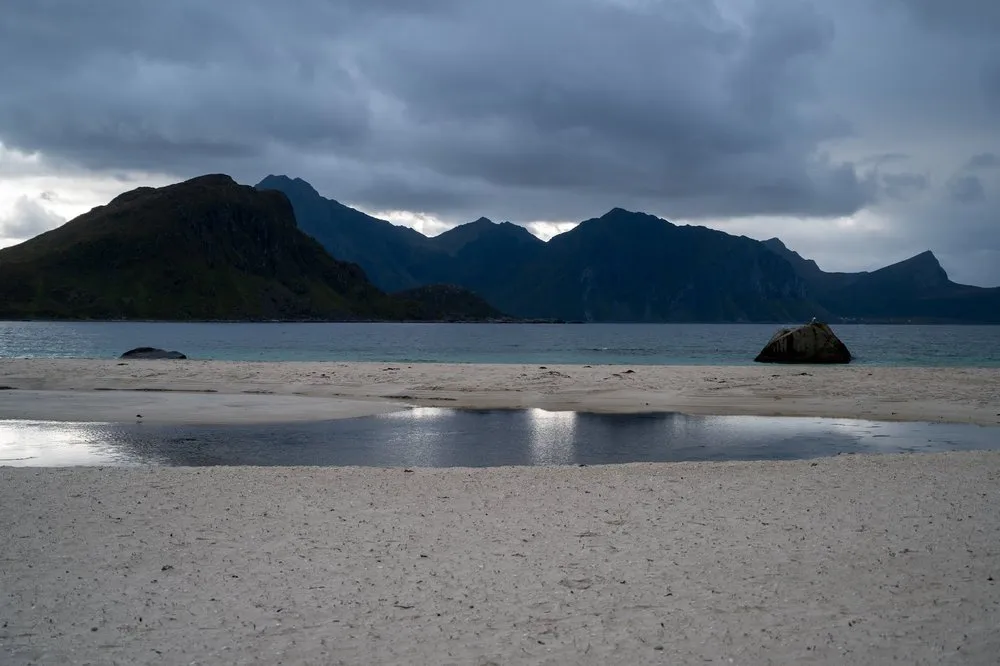 At Hauklandstranda a puddle in the pale sand reflects the sky, with a boulder and dark mountains behind