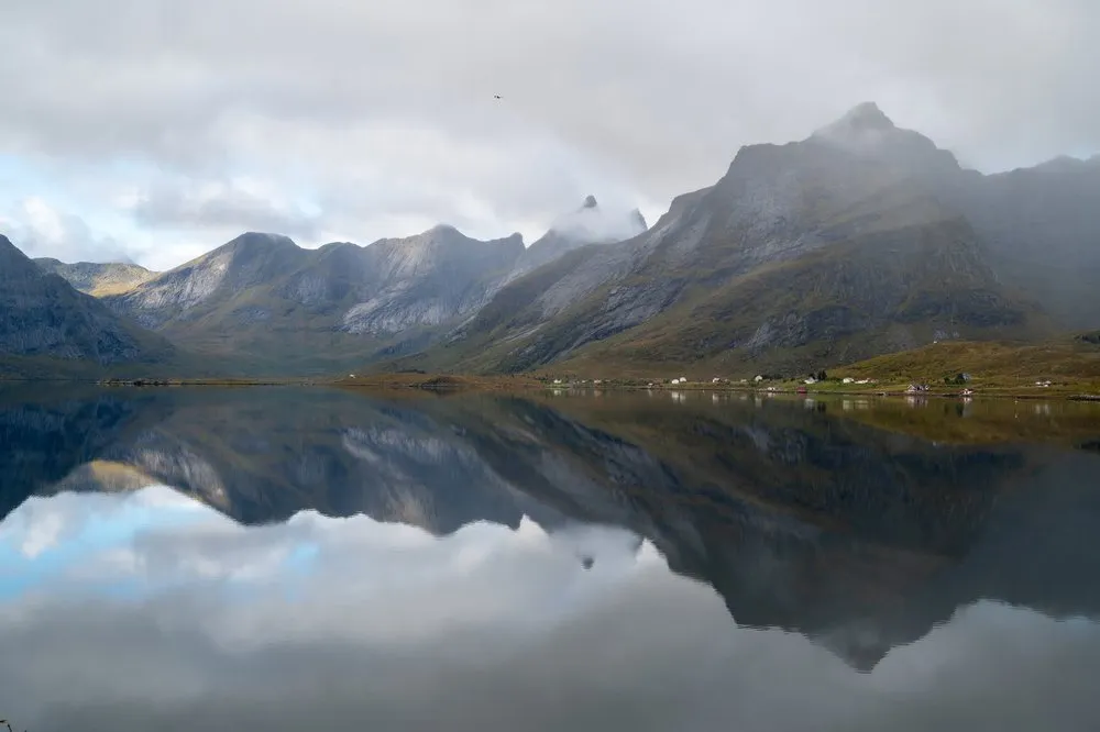 A mountain range beneath clouds is reflected in the smooth sea, scattered houses on the shore