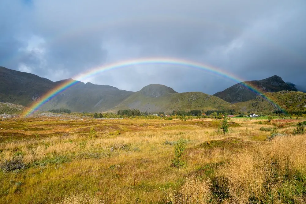 A rainbow arches over a yellow autumn meadow and dark hills, a white house on the right