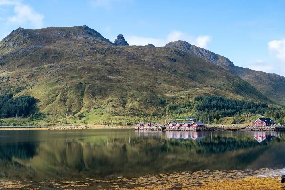 Red huts on the shore are reflected in the calm water, behind them a green mountain with conifer forest under a blue sky