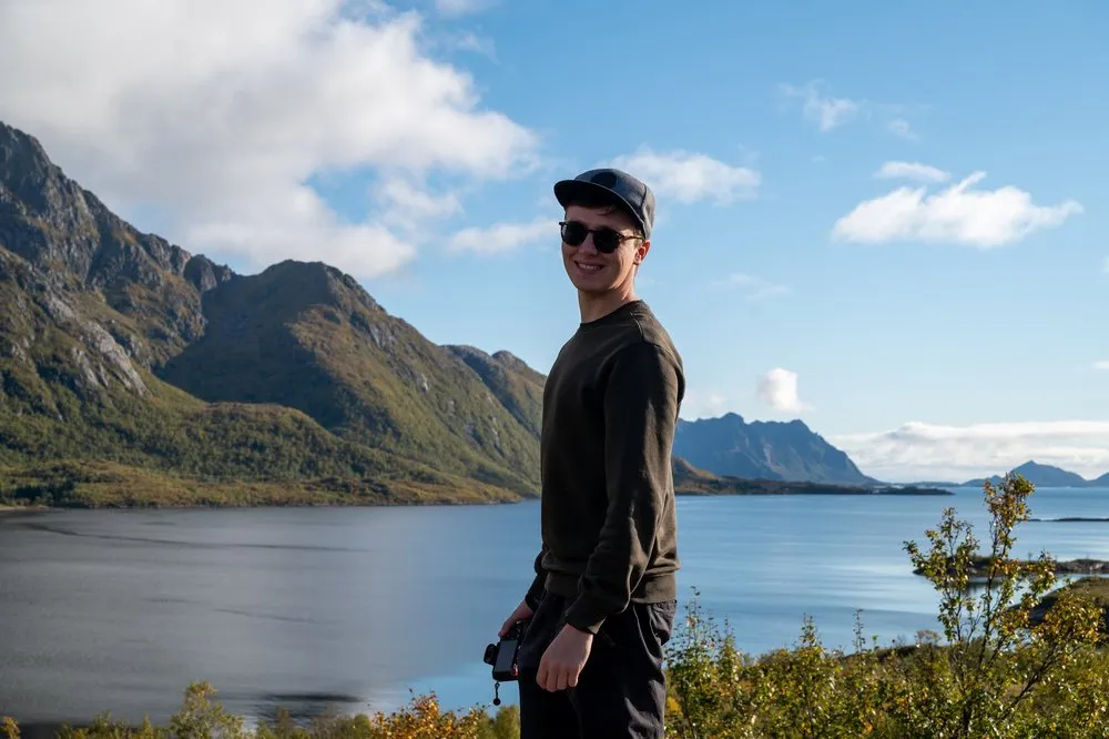 A man in a cap and sunglasses holds a camera, behind him a fjord, mountains and blue sky