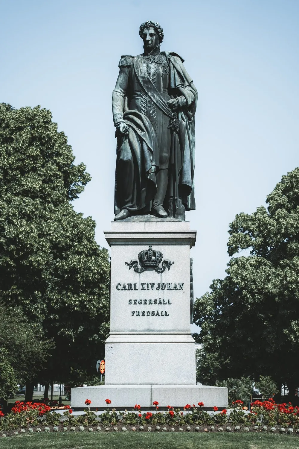 Bronzestatue mit der Inschrift Carl XIV Johan auf einem Steinsockel, umgeben von Bäumen und roten Blumen