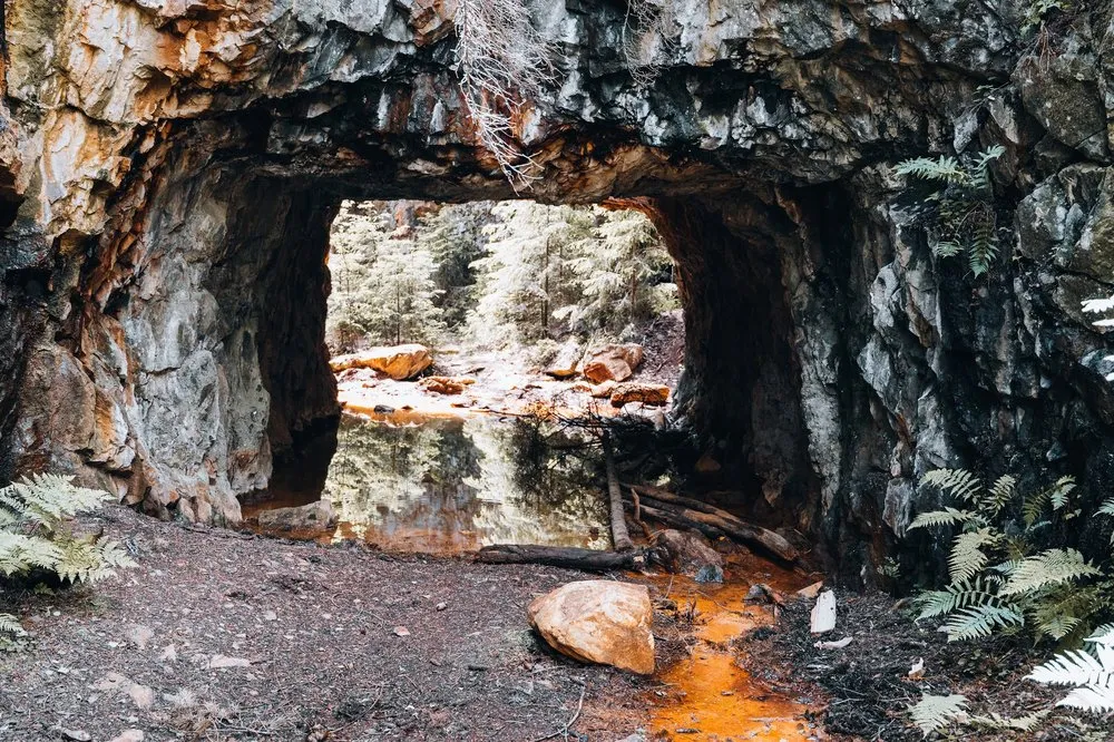Felsdurchgang am Eingang der Mine von Sätragruvan, am Boden orange gefärbtes Wasser, rechts Farn, dahinter Wald