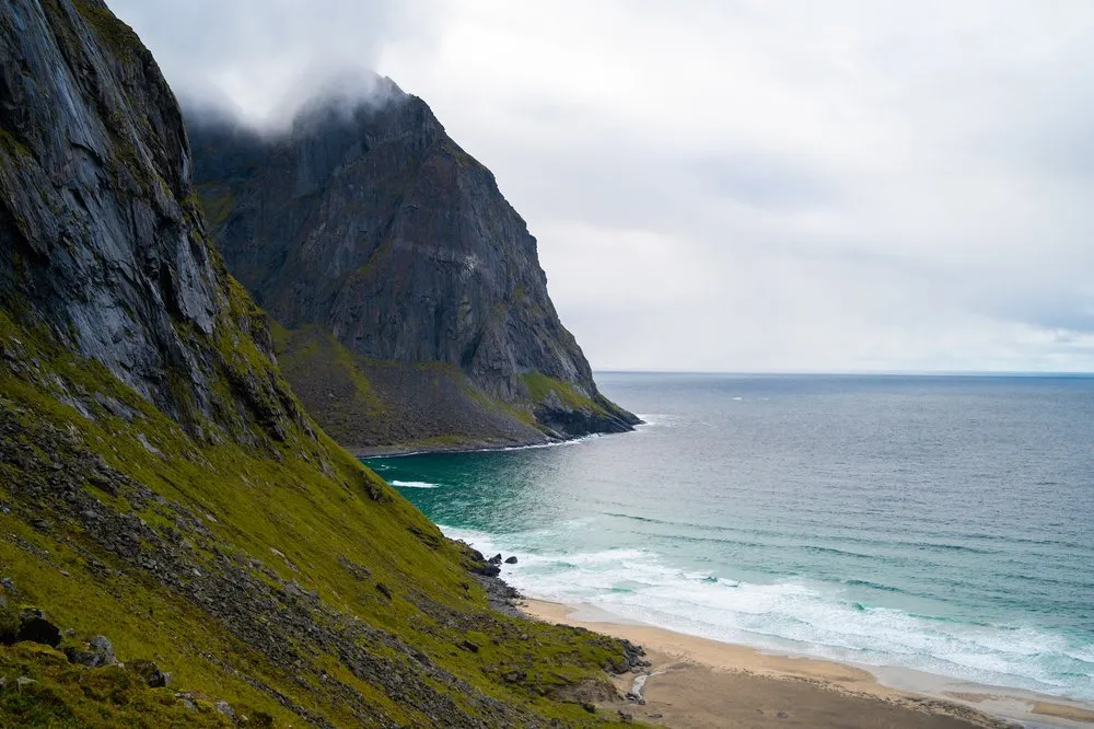 Nebelfetzen an einer steilen Felswand über einem Sandstrand mit türkisem Meer