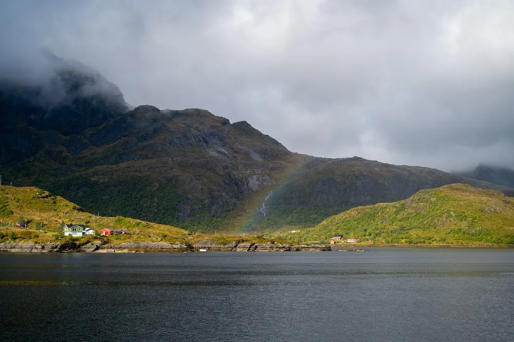 Ein Regenbogen vor einer nebligen Bergflanke, davor ein dunkler Fjord und kleine Häuser am Ufer