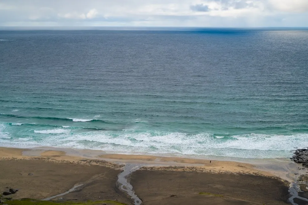 Steiler Blick auf Brandung, Sandstrand und einen Bach, der sich über den Strand windet