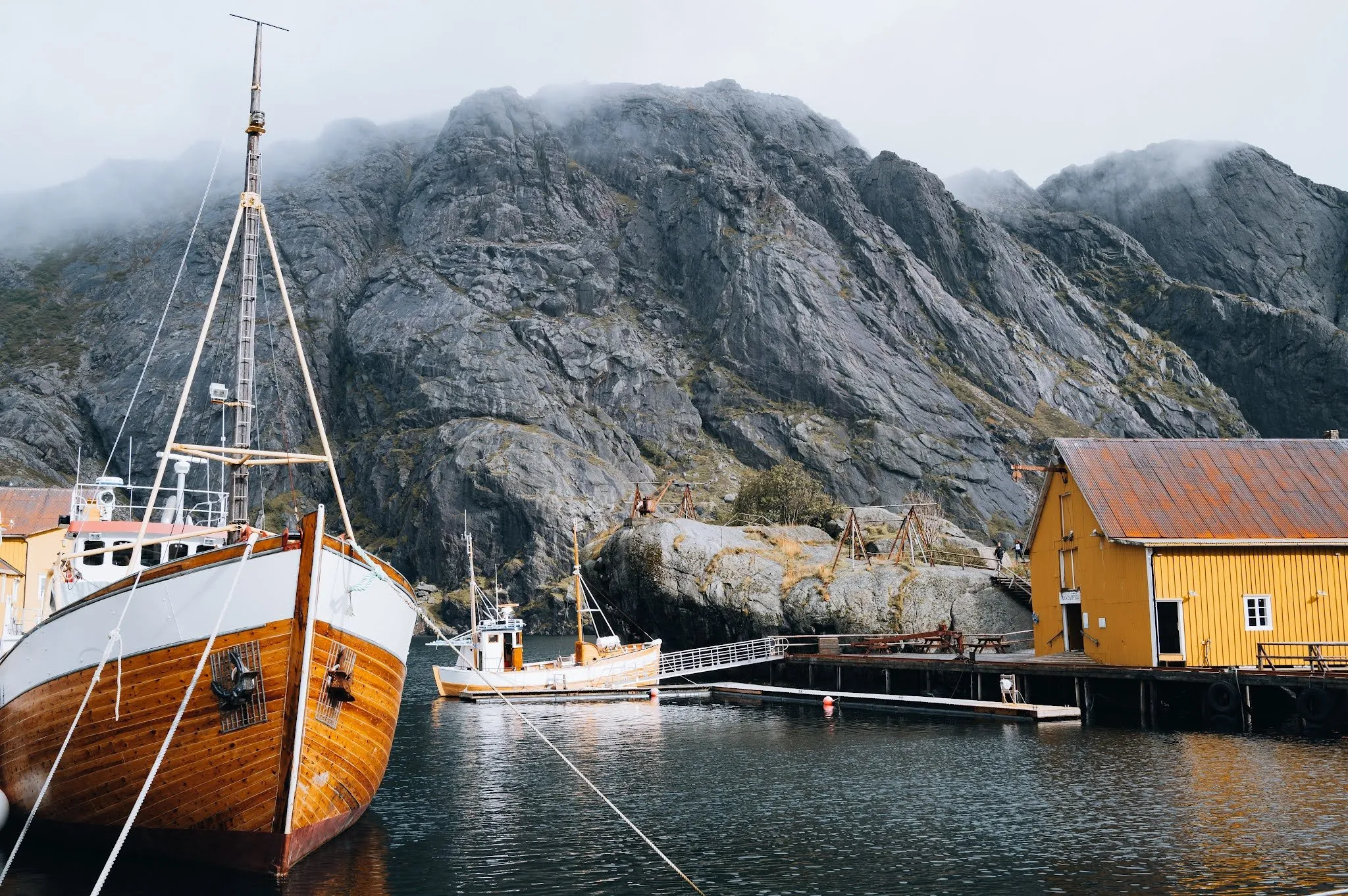 A wooden sailing boat lies in the small harbour next to a yellow wooden house, a grey rock face rising into the fog behind it