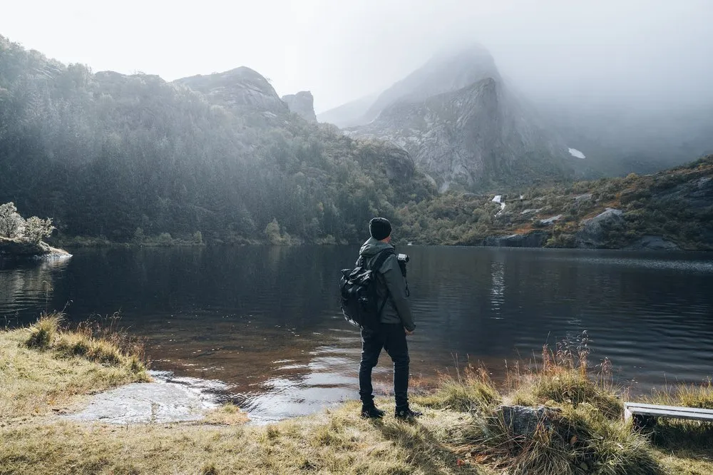 Eine Person mit Rucksack steht am Ufer eines Bergsees, dahinter steigen Berge im Nebel auf