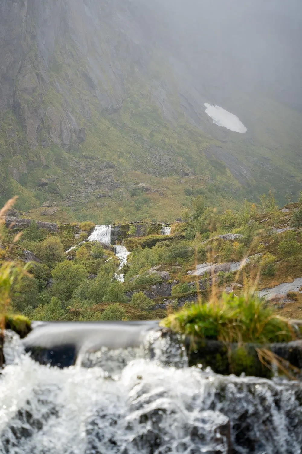Ein Bach stürzt über Felsstufen, dahinter ein Berghang mit kleinen Wasserfällen und einem Schneefeld im Nebel