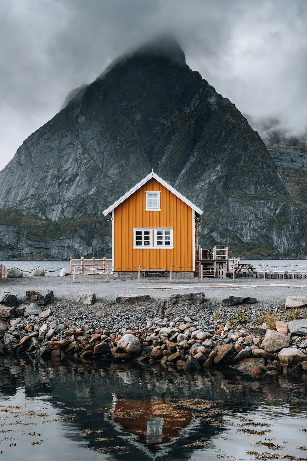 Das gelbe Holzhaus in Sakrisøy vor einem steilen, nebelverhangenen Berg, davor Steine und das spiegelnde Wasser