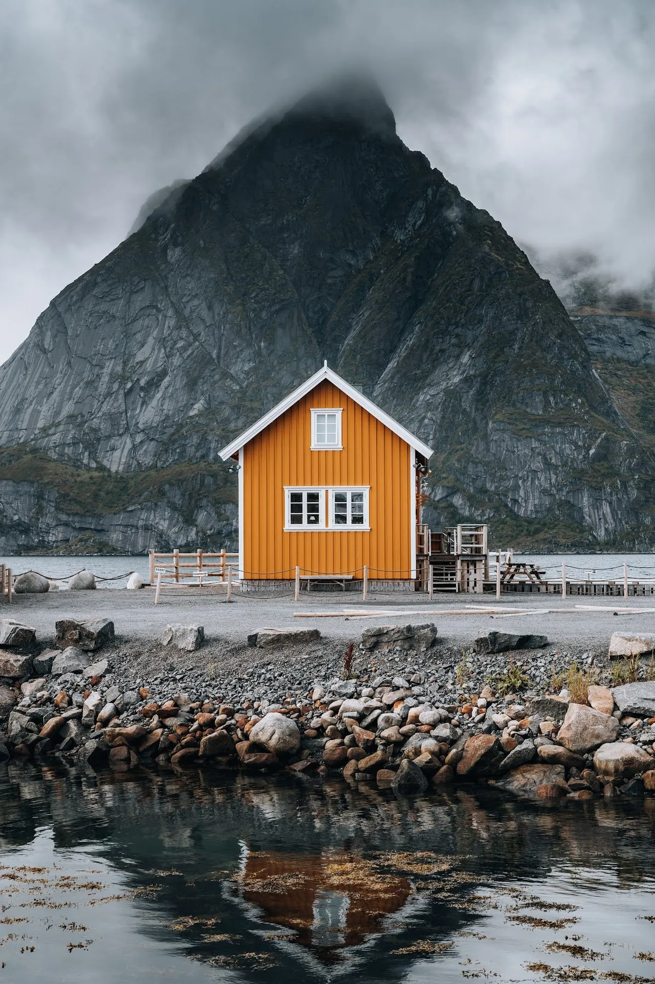 The yellow wooden house in Sakrisøy in front of a steep, fog-shrouded mountain, stones and reflecting water in front of it
