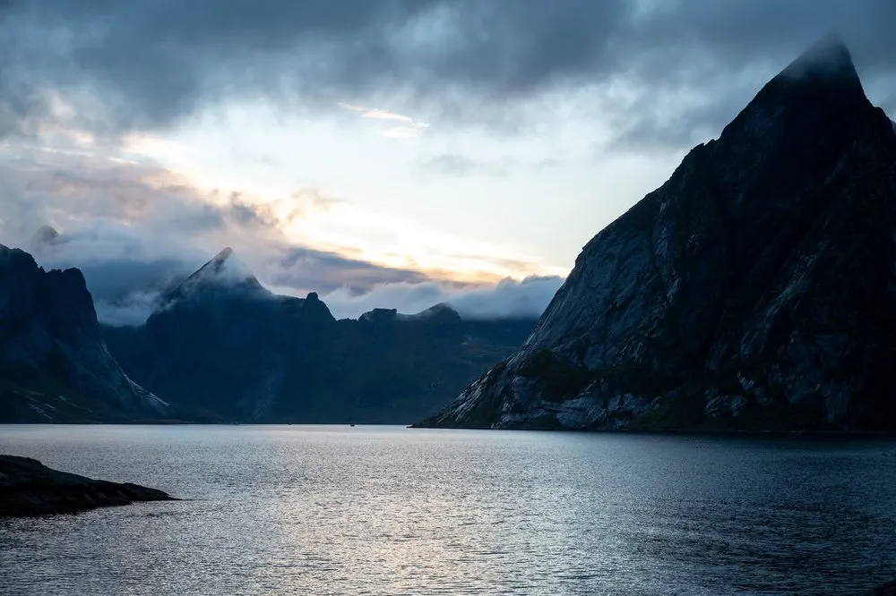 Abendlicht bricht durch Wolken über einem Fjord, rechts ragt ein dunkler Berggipfel auf