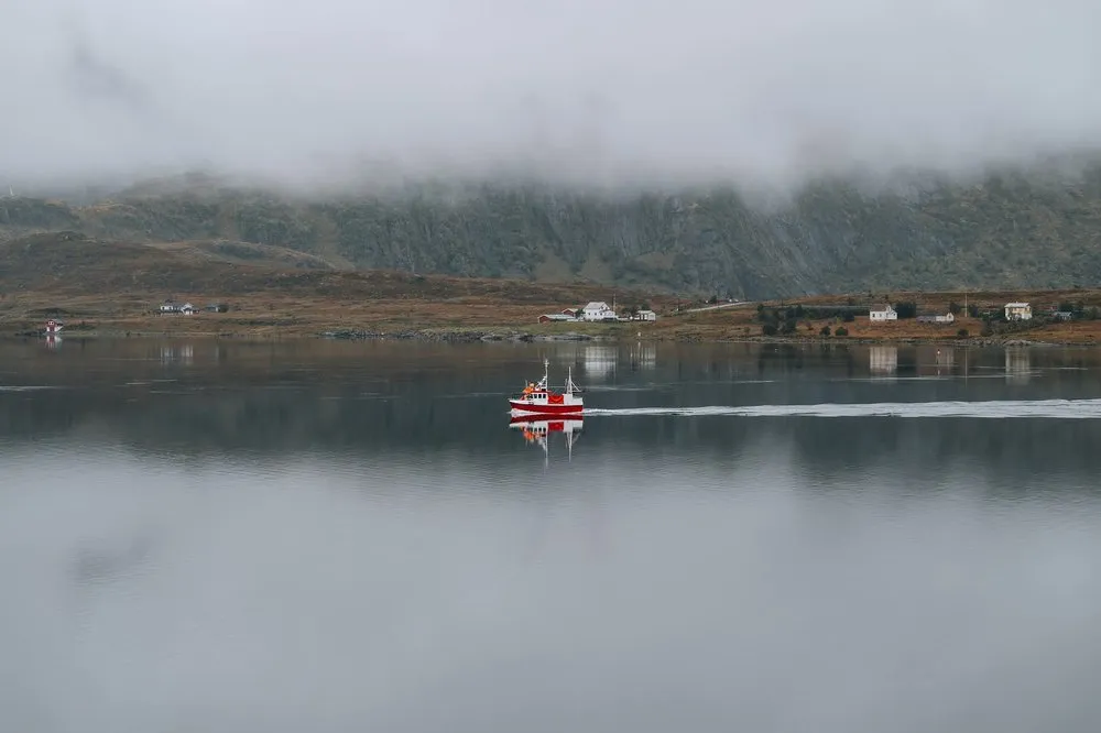 Ein rotes Boot mit weissem Kielwasser fährt über spiegelglattes Wasser, am Ufer stehen einzelne Häuser unter tiefen Wolken