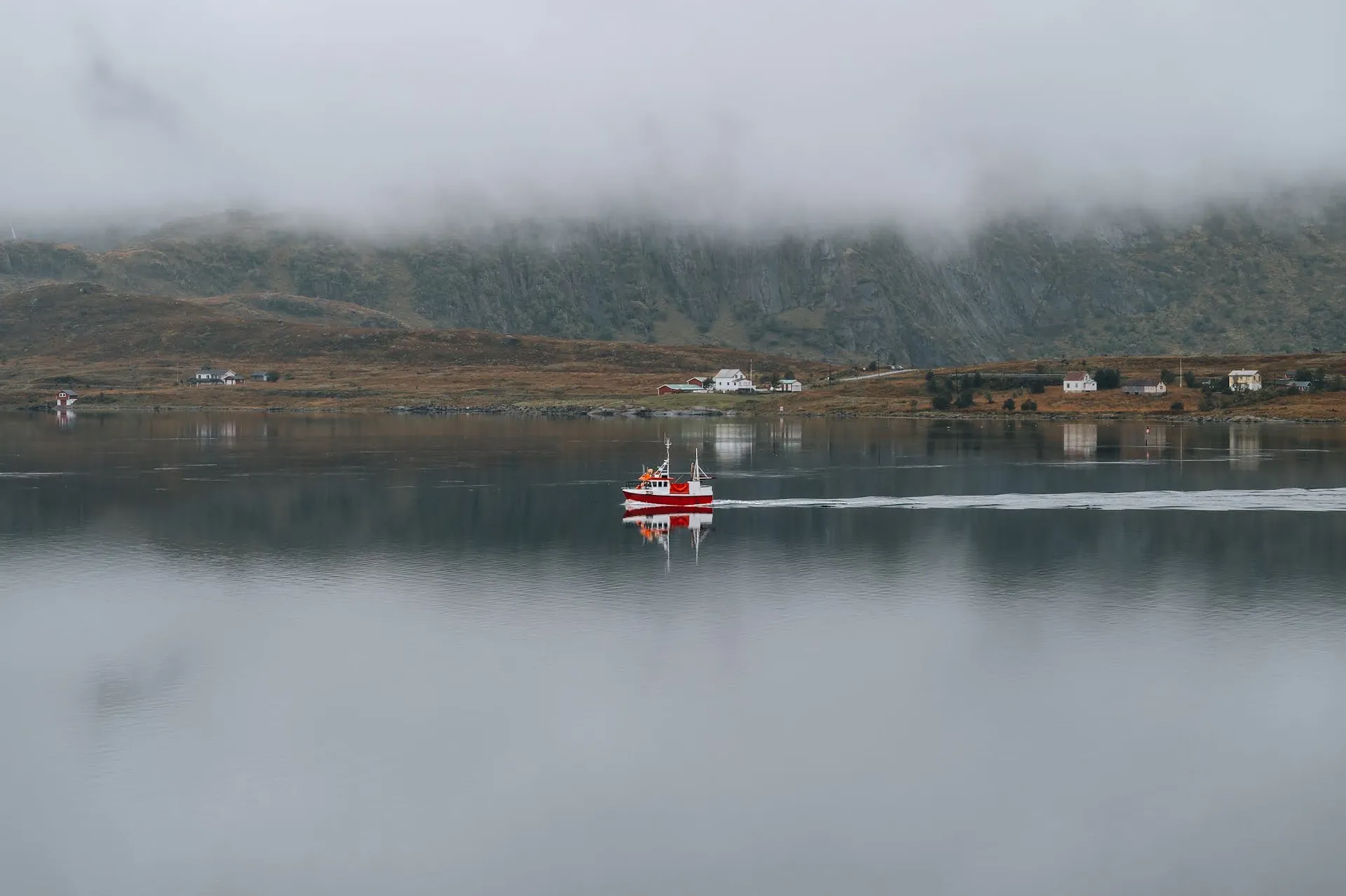 A red boat with a white wake moves across mirror-smooth water, a few houses on the shore under low clouds
