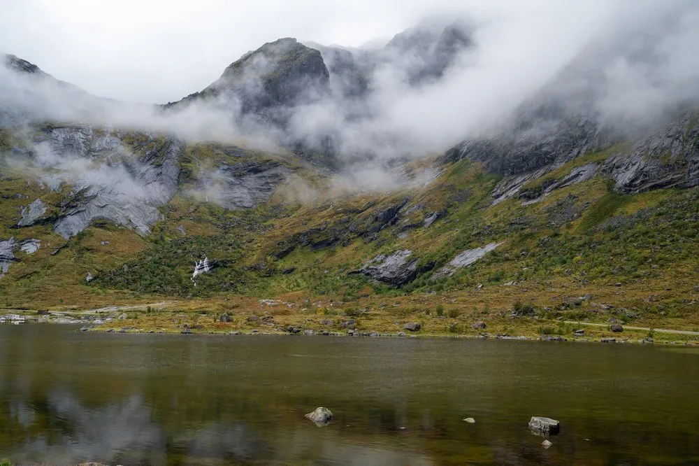 Nebel zieht über einen Berghang mit kleinen Wasserfällen, davor liegt ein dunkler See mit Steinen