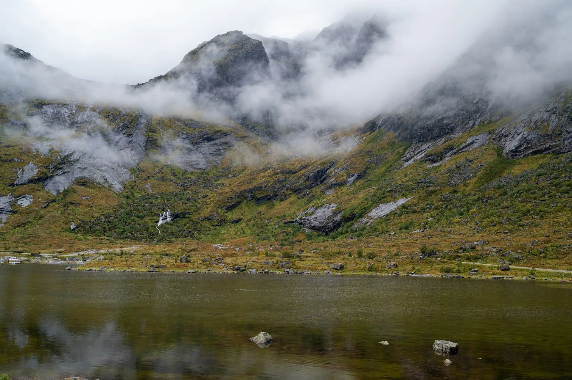 Fog drifts over a mountain slope with small waterfalls, a dark lake with stones in front of it