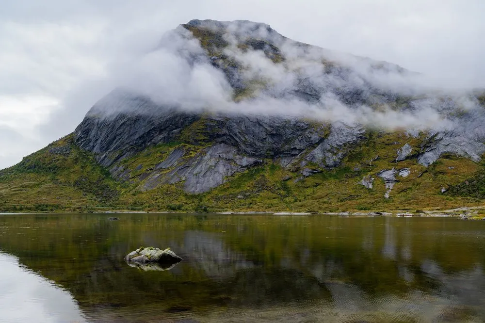 Ein Berg mit einem Wolkenband am Gipfel spiegelt sich im See, im Vordergrund ein bemooster Stein