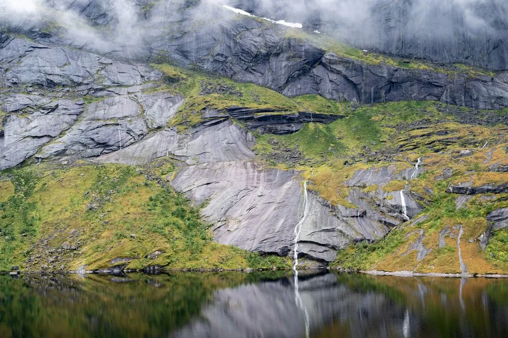 Eine graue Felswand mit grünen und gelben Flecken, an der mehrere schmale Wasserfälle herabfallen, spiegelt sich im See