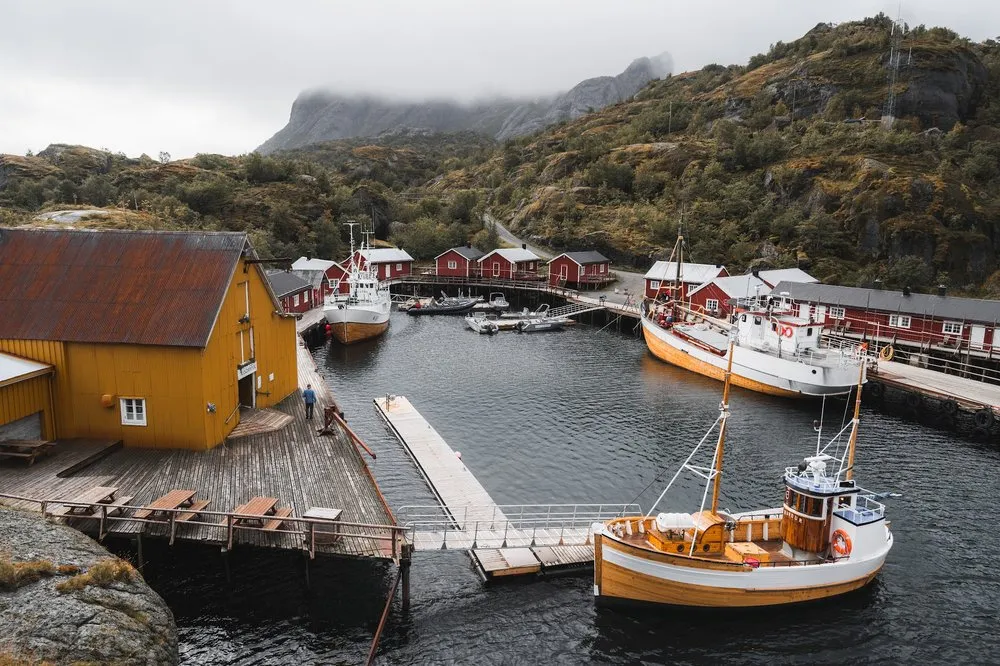 Blick von oben auf den Hafen von Nusfjord mit einem gelben Holzhaus, roten Rorbu's und mehreren Holzbooten