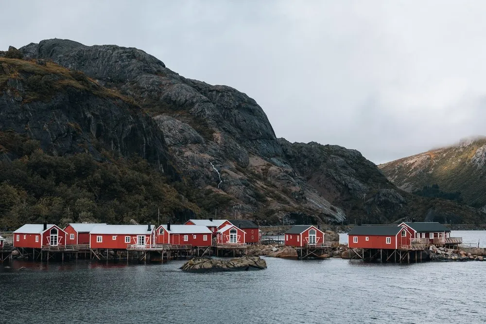 Eine Reihe roter Rorbu's auf Stelzen am Wasser in Nusfjord, dahinter ein dunkler Felshang