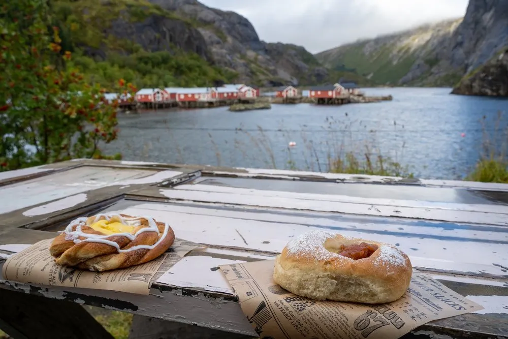 Zwei Gebäckstücke auf Zeitungspapier auf einem alten Holztisch, dahinter das Wasser von Nusfjord mit roten Rorbu's
