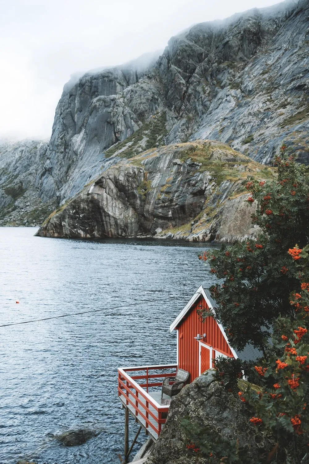Eine rote Holzhütte mit Terrasse auf Stelzen steht über dem Wasser vor einer steilen Felswand, daneben ein Baum mit orangen Beeren