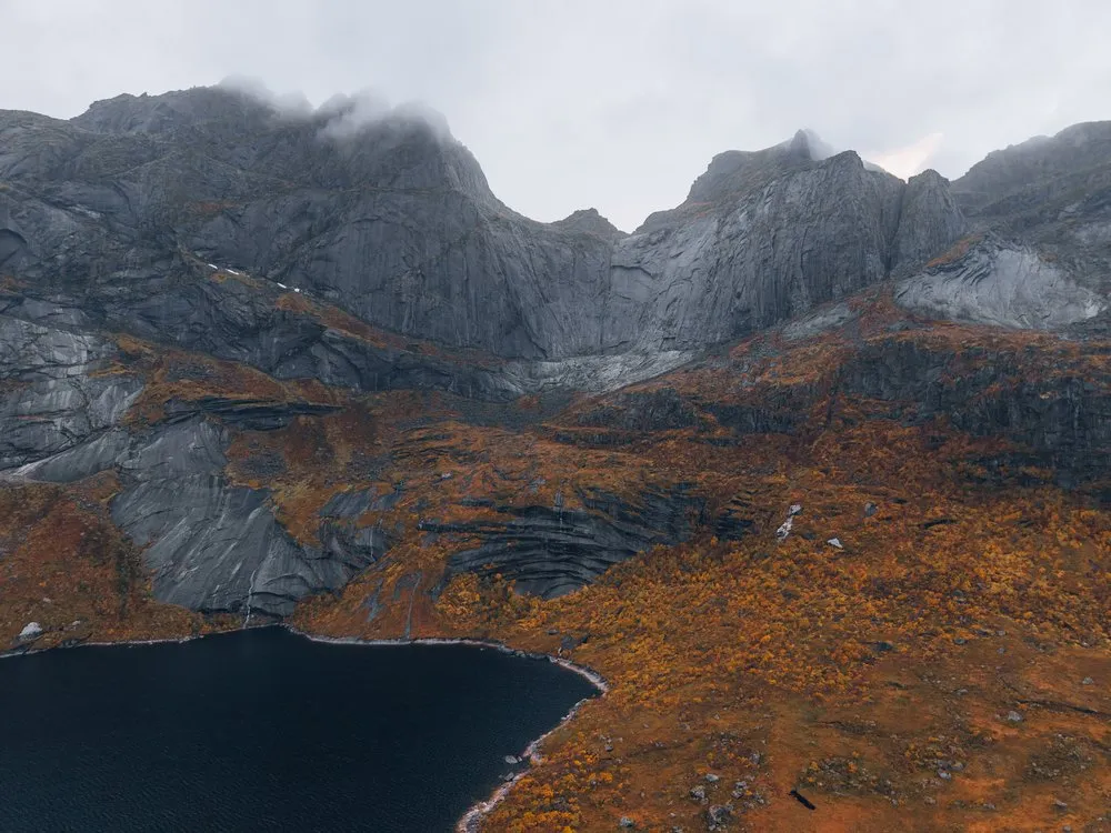Graue Felswände mit Wolken an den Gipfeln, unten ein dunkler See und orange Hänge an der Strasse nach Nusfjord