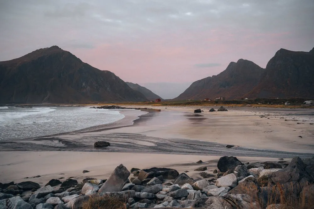 Der Flakstadstranda im Abendlicht, vorne Steine, dahinter der Sandstrand und Berge unter rötlichem Himmel