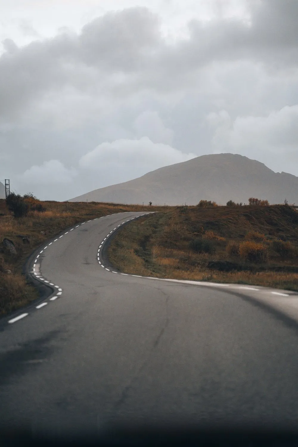 Eine kurvige Strasse zwischen braunen Herbstwiesen auf der Fahrt vom Flakstadstranda nach Ramberg, dahinter ein Berg unter Wolken