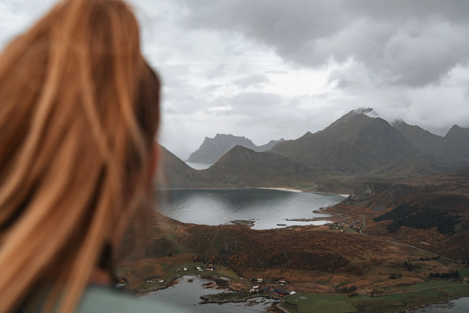 Eine Wanderin mit langen Haaren schaut vom Offersøykammen hinunter auf die Bucht, die Häuser am Ufer und die Berge