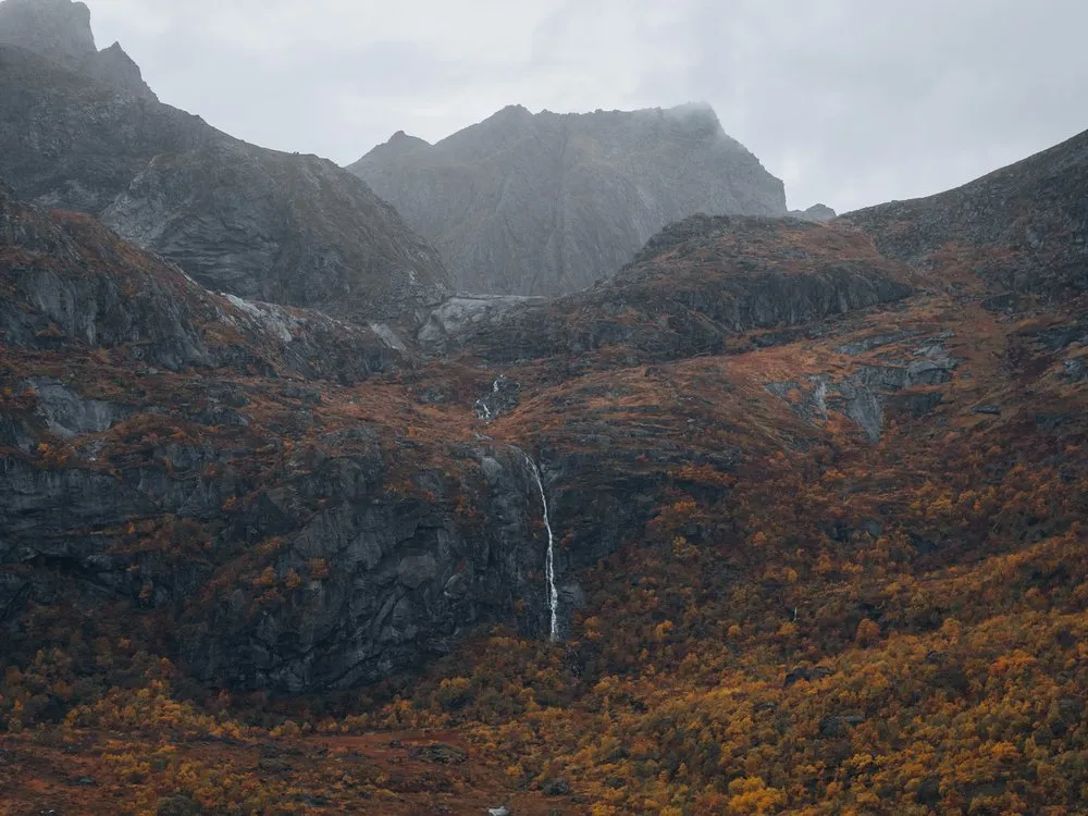 Ein schmaler Wasserfall an einer dunklen Felswand über orange gefärbten Hängen an der Strasse nach Nusfjord