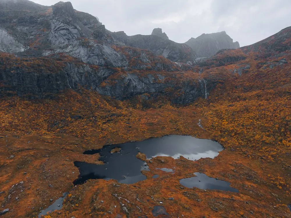 Ein kleiner dunkler See in rotbrauner Herbstlandschaft unter grauen Felswänden bei Nusfjord, aus der Luft