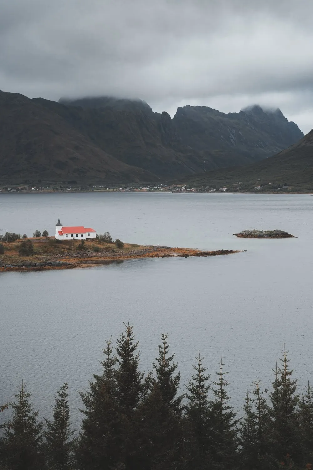 Small white church with a red roof on a spit of land in the water, mountains behind it in dark clouds