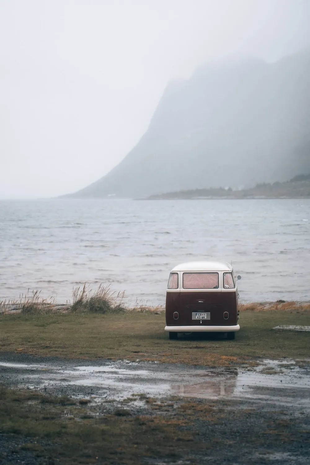 An old VW bus stands on a meadow by the water, fog and a steep mountain slope behind it