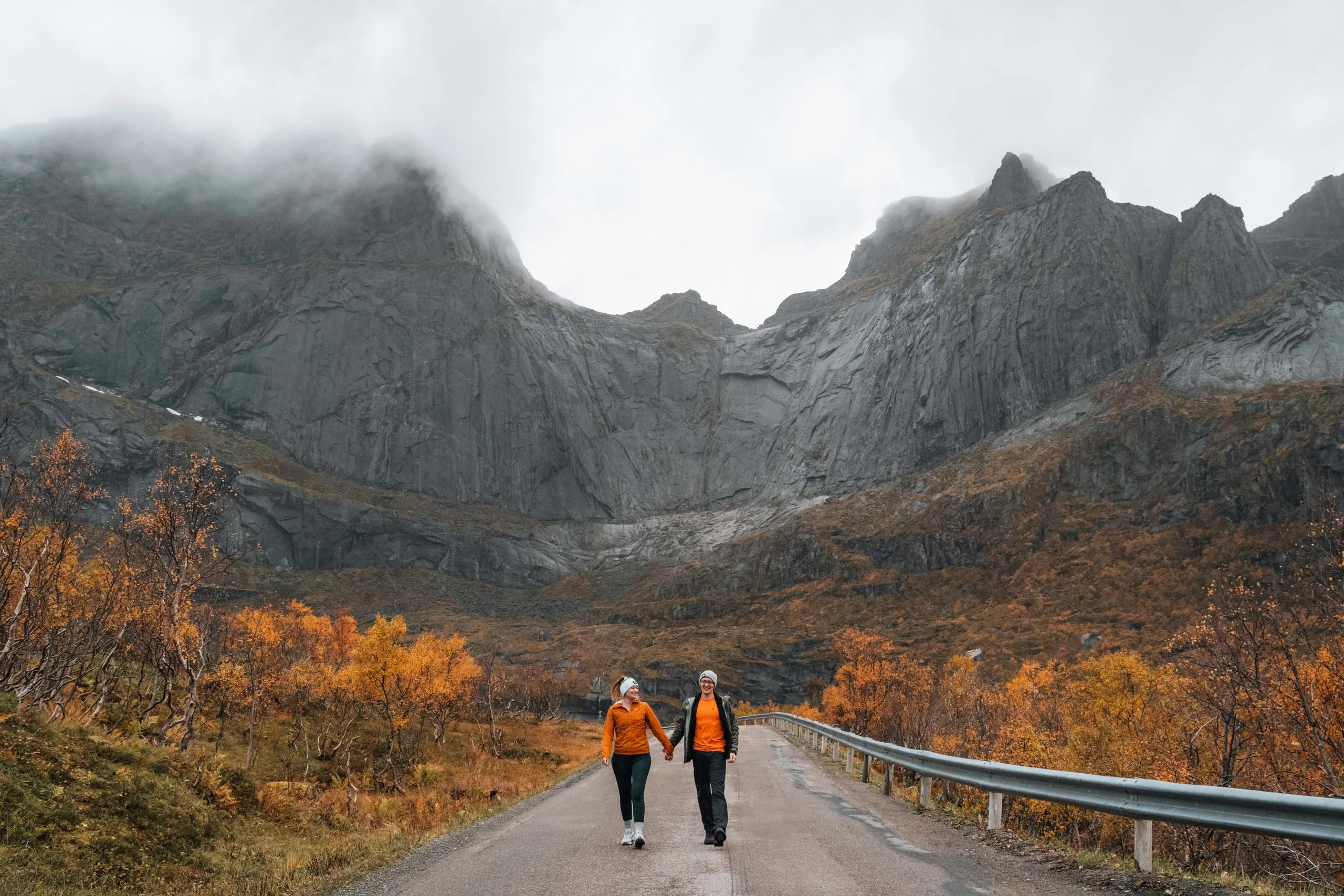 Annina und Nicolas gehen Hand in Hand auf der Strasse nach Nusfjord, beide in orangen Jacken, hinter ihnen graue Felswände im Nebel und herbstlich gefärbte Birken
